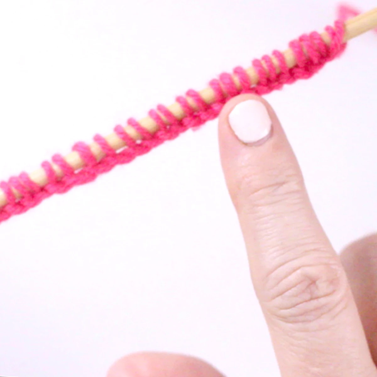 Woman's finger pointing to knitting stitches on a knitting needle in pink color yarn.