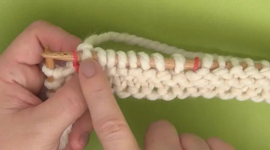 A close up of hands holding a knitting needle with white yarn cast-on stitches