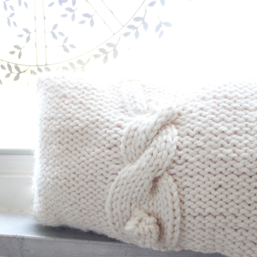 A knitted pillow with Bunny Cable stitch pattern propped up on a window in white yarn