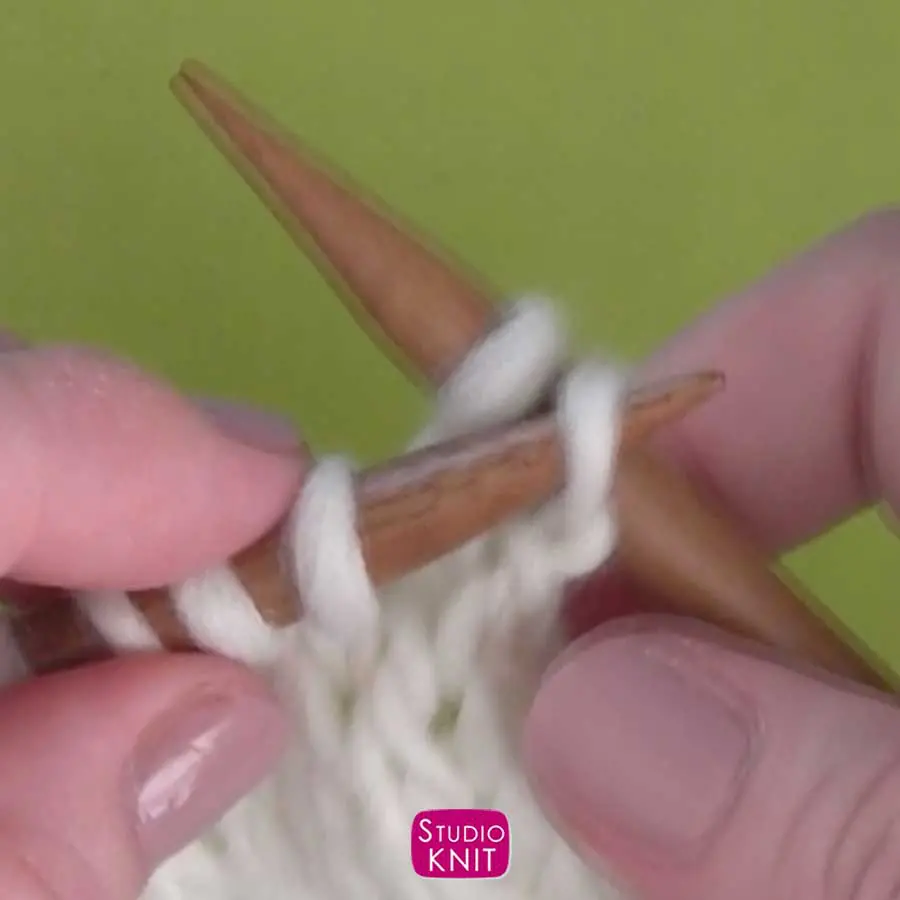 Hands demonstrating how to pick up a stitch on the right knitting needle from the left with white yarn 