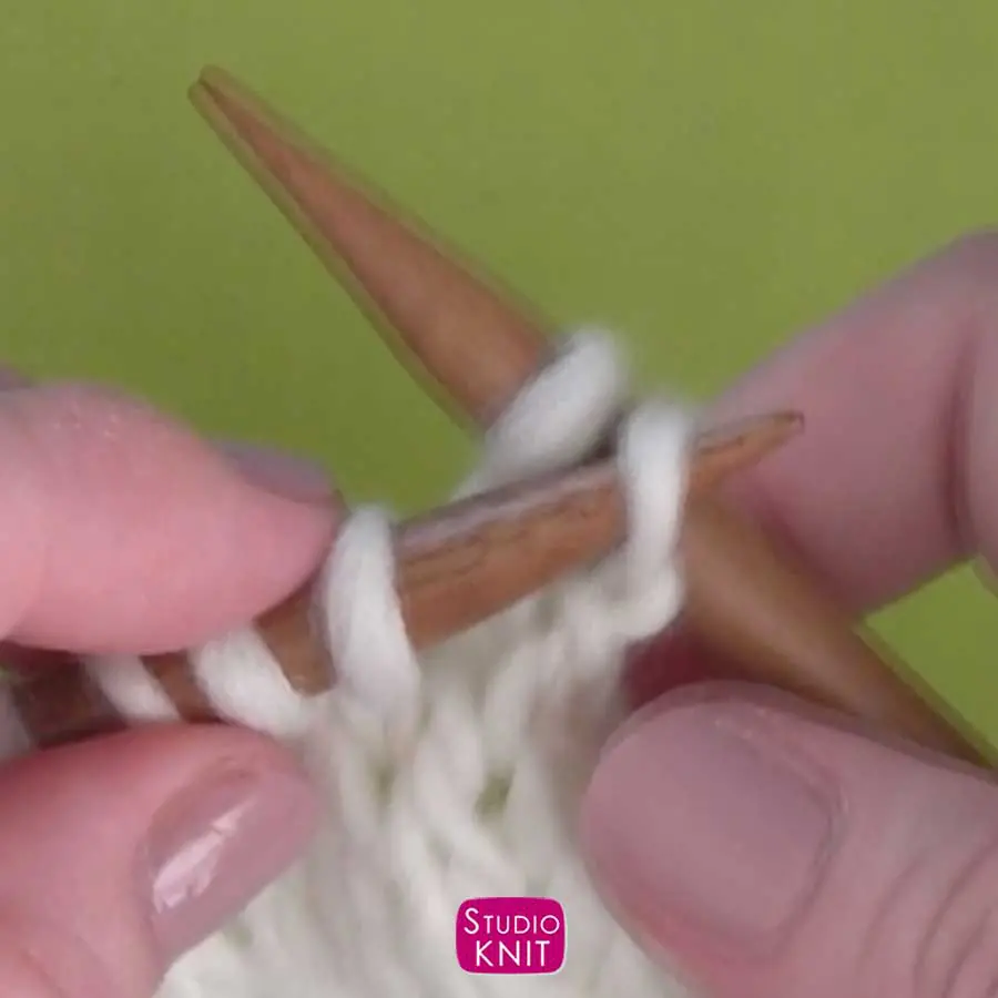 Hands demonstrating how to pick up a stitch on the right knitting needle from the left with white yarn