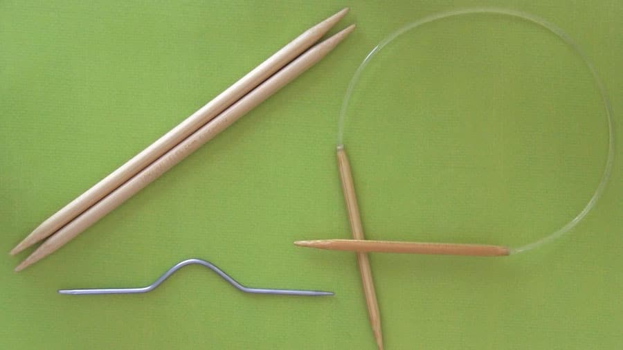 Two straight knitting needles, a circular knitting needle, and a metal cable needle on a green background