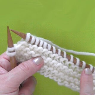 knitted swatch of garter stitch on knitting needles in white color yarn held by hands.