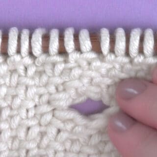 Knitted swatch in white yarn with buttonhole opening on a knitting needle with woman's hand.