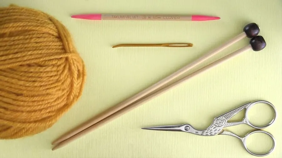Knitting materials with yellow yarn, knitting needles, a tapestry needle, and scissors.