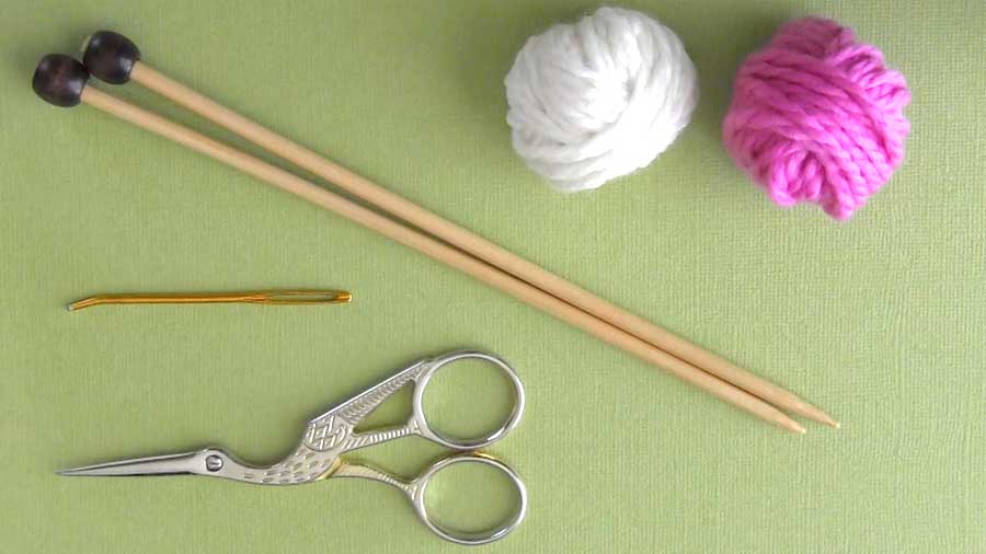 White and pink yarn with knitting needles, scissors, and a tapestry needle.
