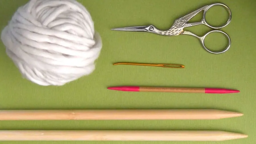 White yarn, stork scissors, tapestry needle, double-pointed knitting needle, and straight knitting needles on a green background