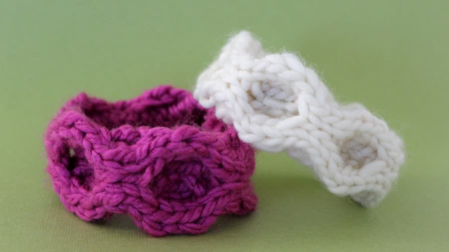 Two knitted bracelets in the honeycomb cable stitch pattern with white and magenta yarn colors on a green background