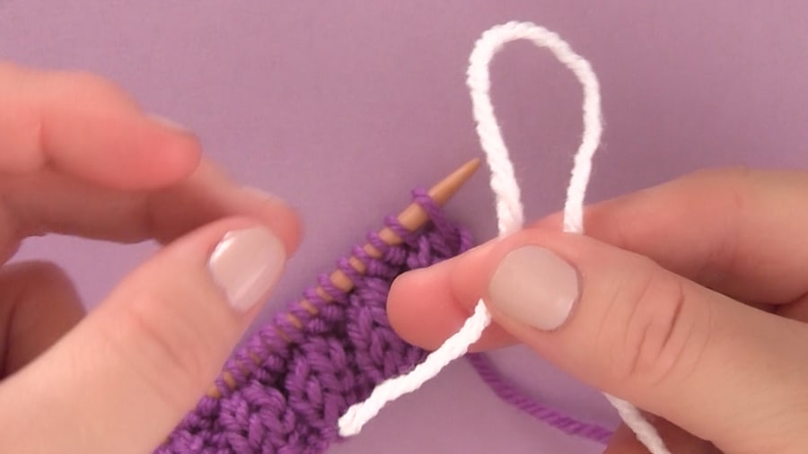 A woman\'s hand holding a strand of white yarn with a purple knitted swatch on a knitting needle.