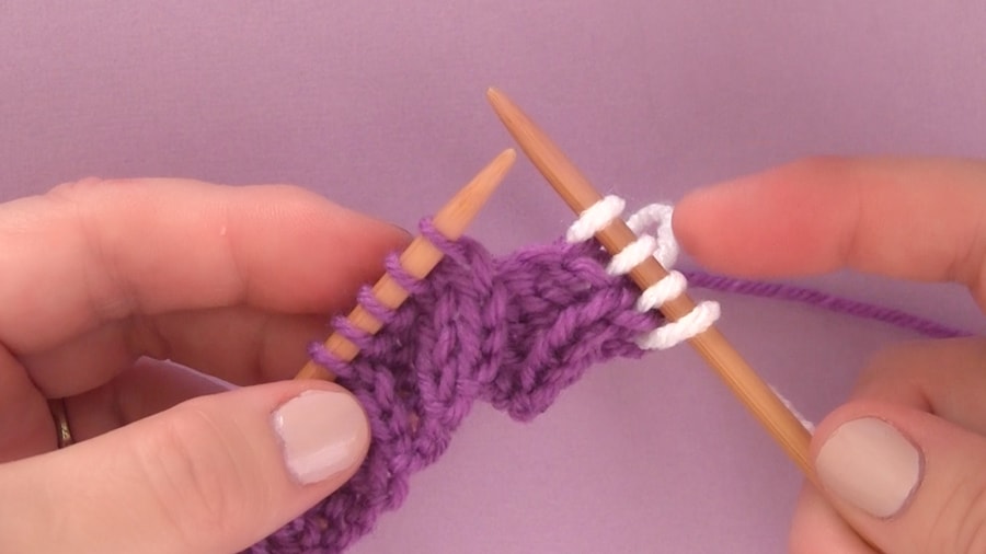 Two straight knitting needles held by a woman\'s hands with purple and white yarn stitches.