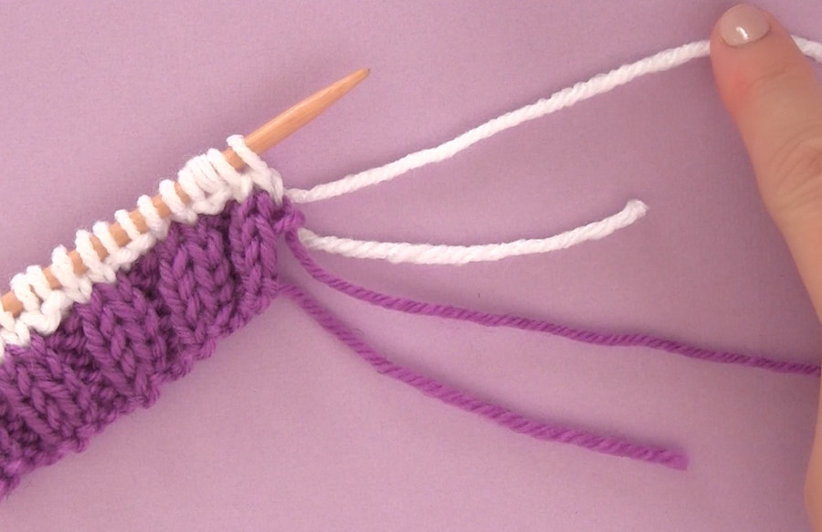 One straight knitting needle with purple and white knitting swatch atop a lavender background.
