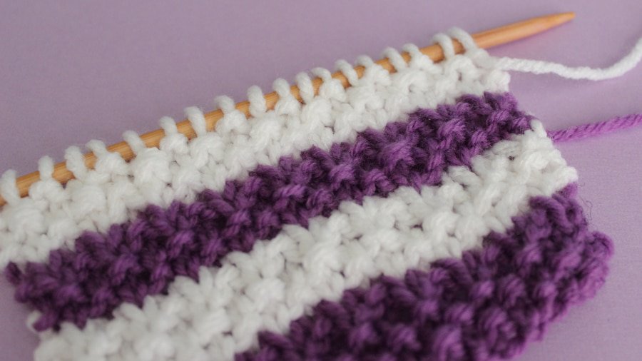Knitted swatch of purple and white yarn on a straight knitting needle.