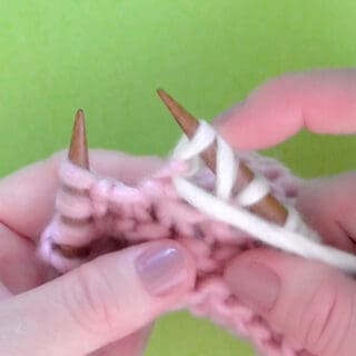 Woman's hands holding knitting needles demonstrating the yarn over knitting technique in pink and white color yarn.
