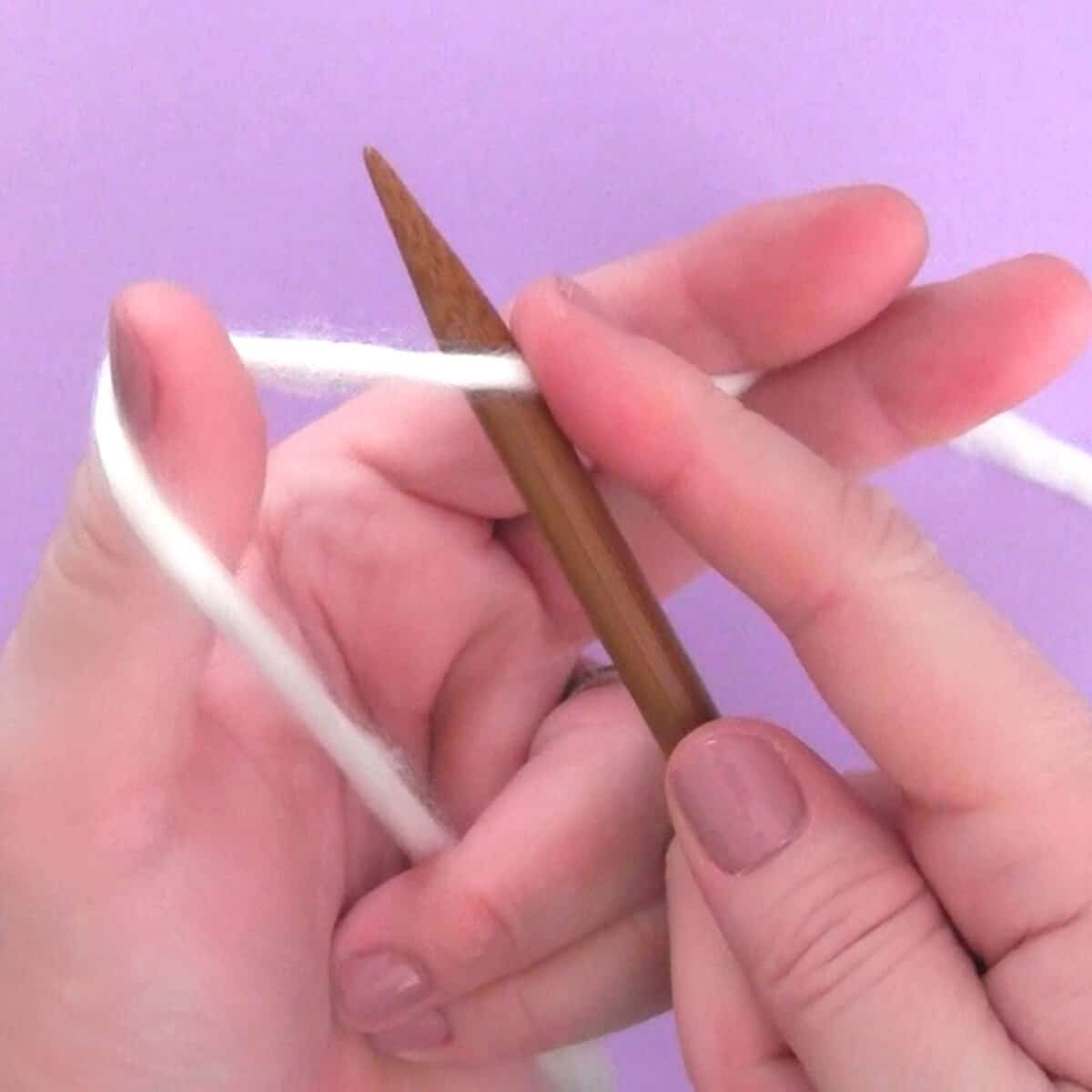 Hands demonstrating how to cast on without a slip knot with white yarn and wooden bamboo knitting needles atop a purple background.