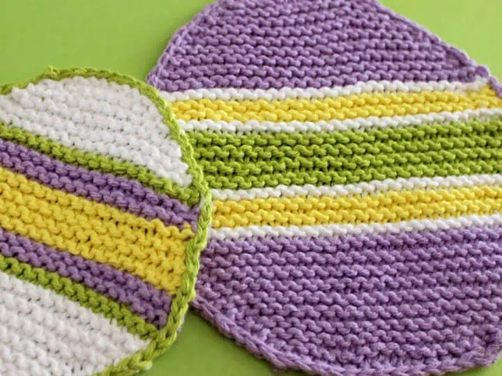 Two knitted Easter Egg Dishcloths in purple, white, yellow, and green stripes atop green backdrop.