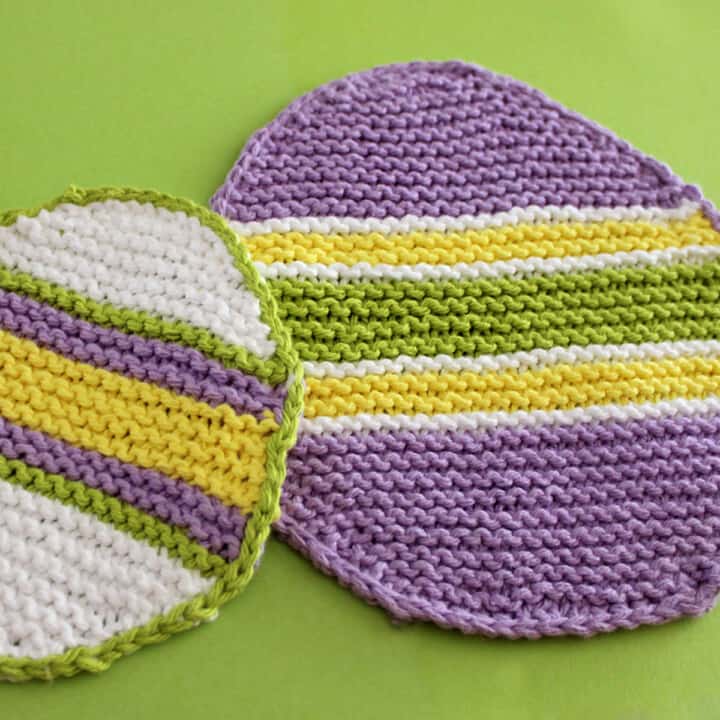 Two knitted Easter Egg Dishcloths in purple, white, yellow, and green stripes atop green backdrop.