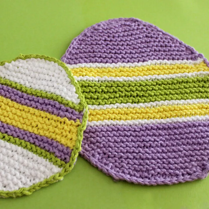 Two knitted Easter Egg Dishcloths in purple, white, yellow, and green stripes atop green backdrop.