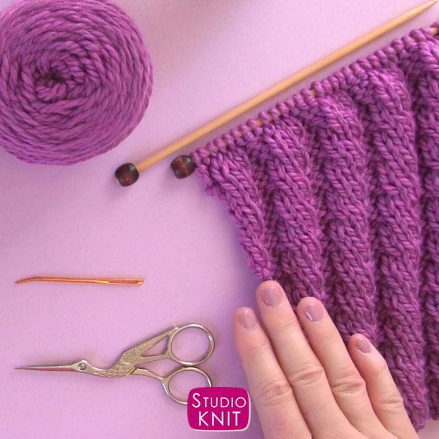 knitting materials with purple yarn, knitting needles, scissors, and a tapestry needle