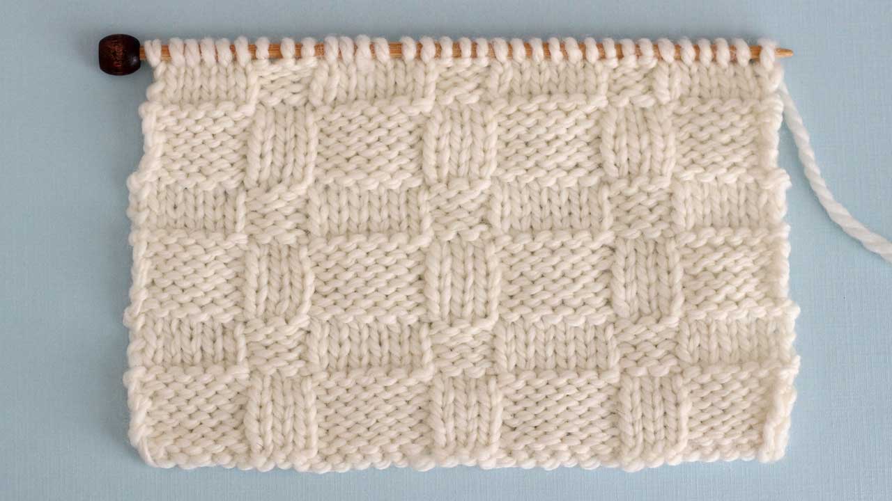 Close up a swatch of textured Wide Basket Weave Stich in white yarn on a knitting needle