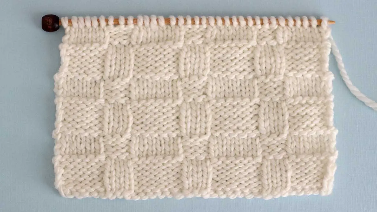 Close up a swatch of textured Wide Basket Weave Stich in white yarn on a knitting needle