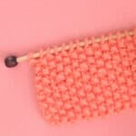 Seed Stitch Knitting swatch on a knitting needle in orange yarn.