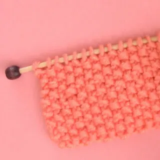 Seed Stitch Knitting swatch on a knitting needle in orange yarn.