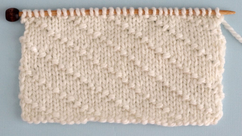 Swatch of textured Diagonal Seed Stitch swatch in white yarn on a knitting needle