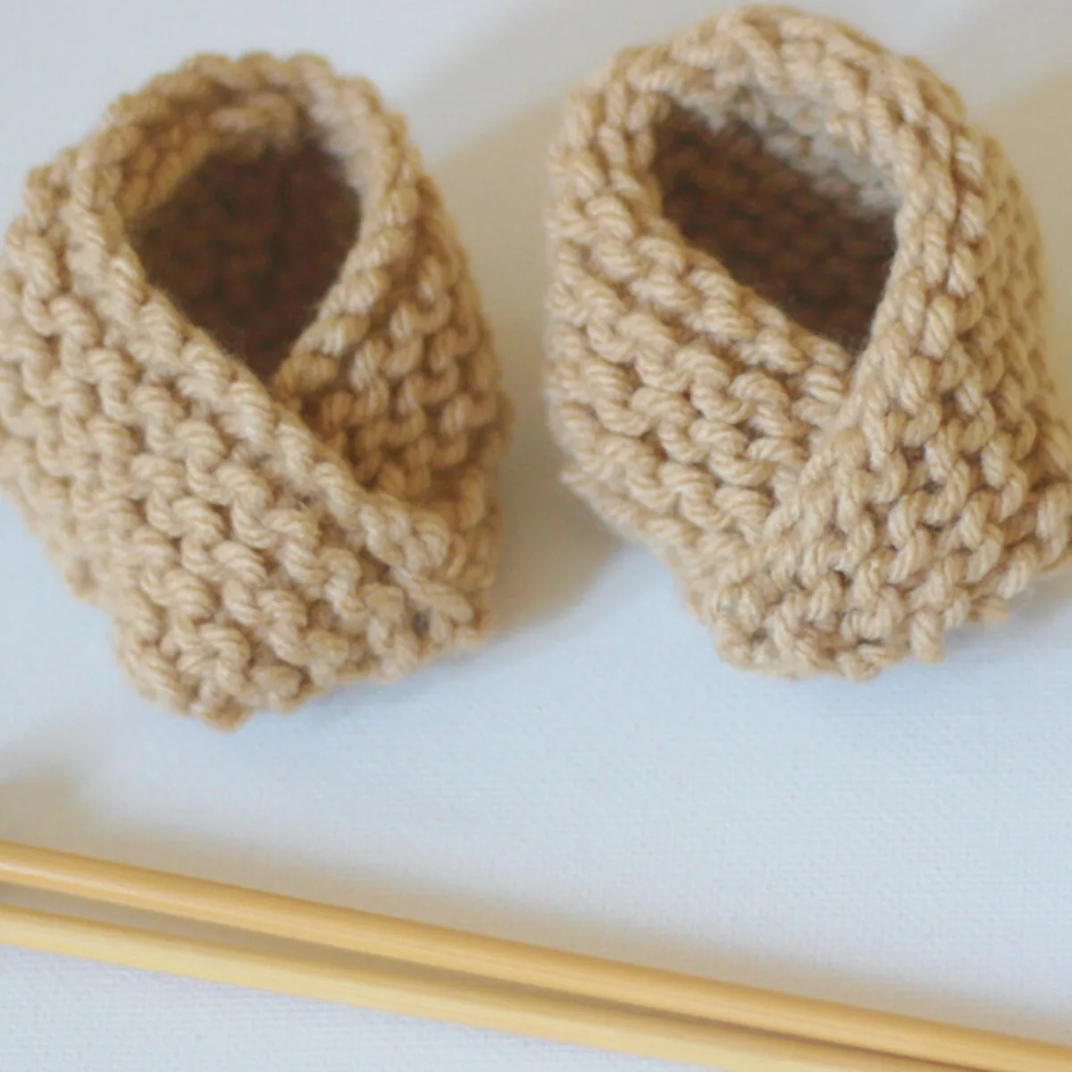 Knitted baby booties in garter stitch with beige yarn color and knitting needles.