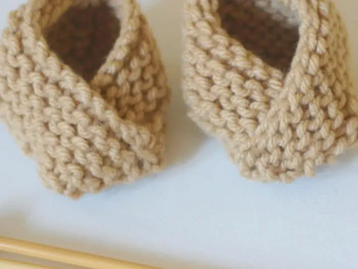 Knitted baby booties in garter stitch with beige yarn color and knitting needles.