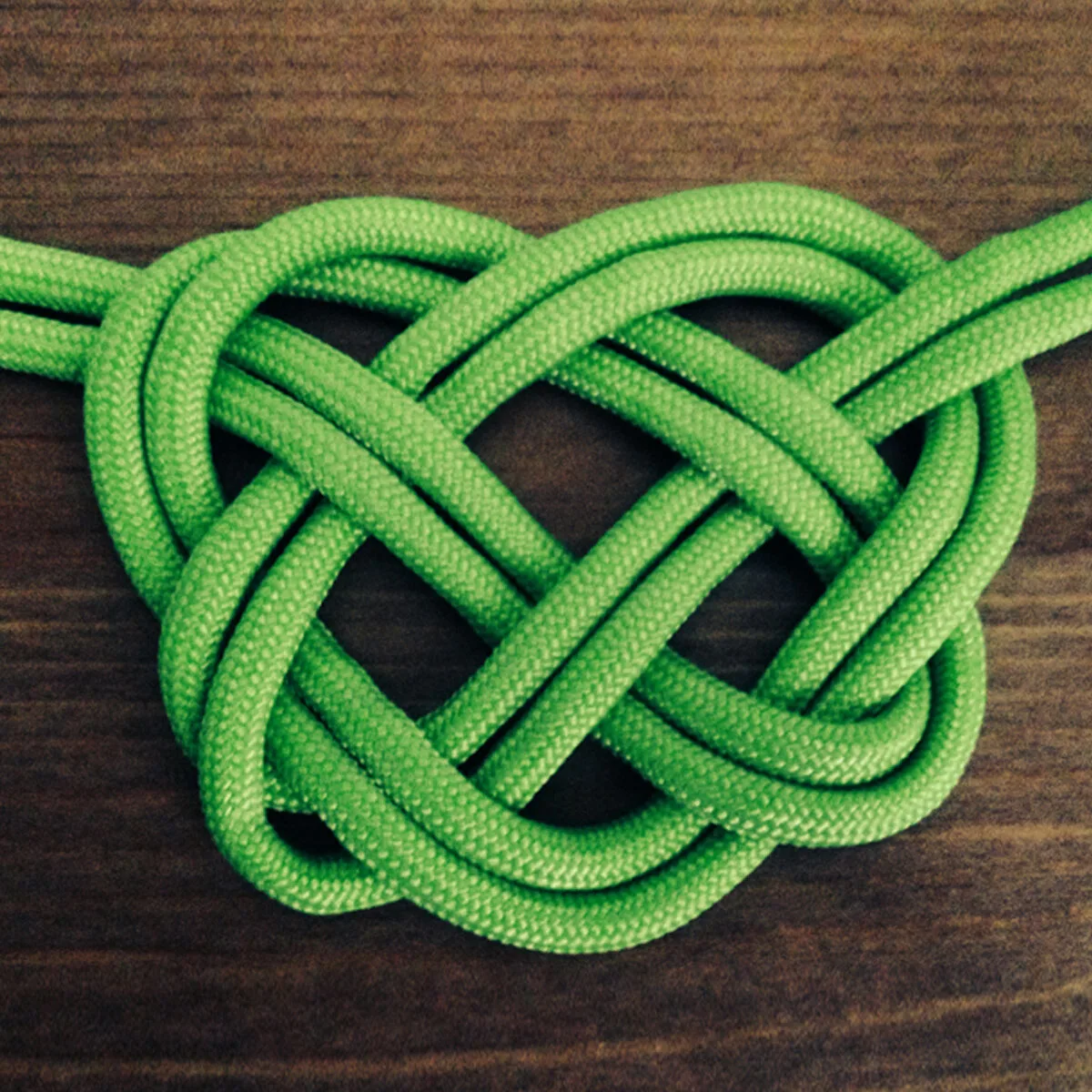 Celtic Heart Knot with double strands of green colored rope atop wooden background.