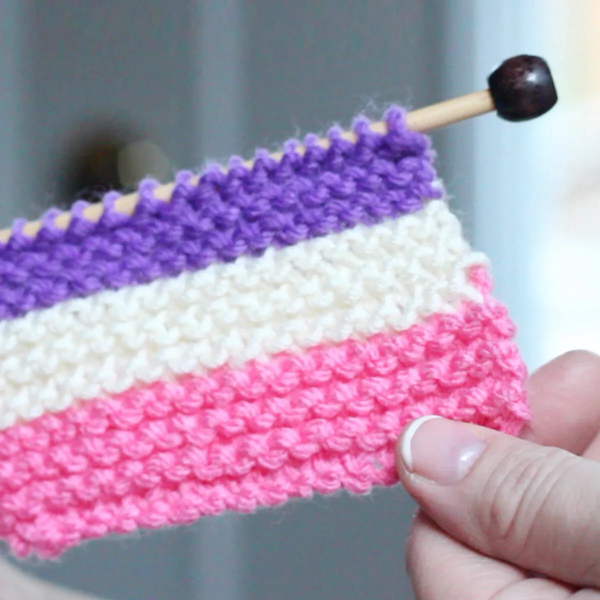 Knitted Swatch of horizontal stripes in purple, white, and pink color yarn on knitting needle with hands holding.