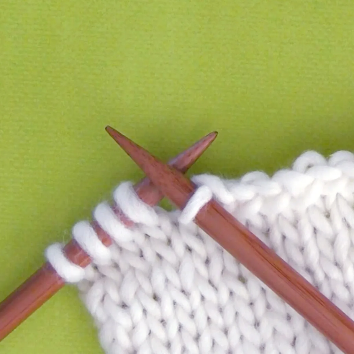 Knitted swatch of white yarn in stockinette stitch pattern with two wooden knitting needles.