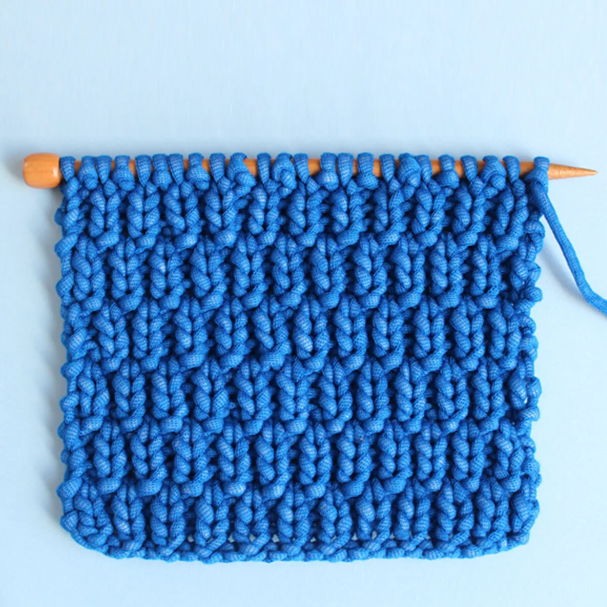 Long Raindrops Knit Stitch Pattern in bright blue yarn color on knitting needle atop a light blue background.
