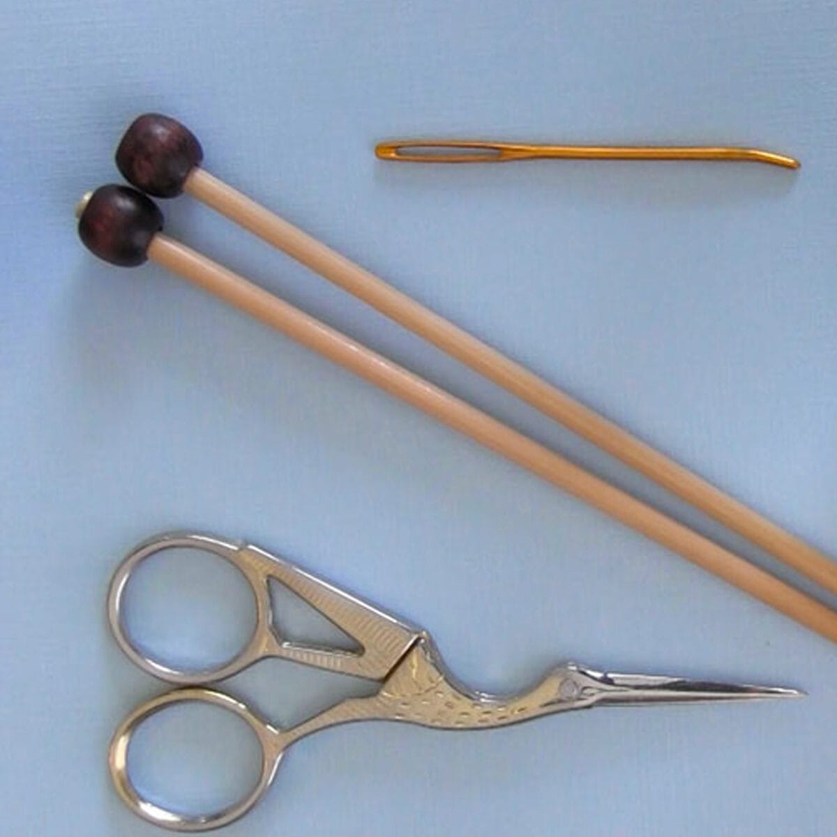 Two straight wooden bamboo knitting needles, a pair of stork shaped scissors, and a tapestry needle atop a blue background.