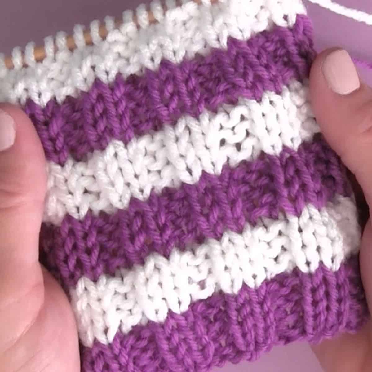Knitted horizontal stripe pattern in rib stitch texture with purple and white yarn colors held by hands.