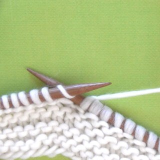Two straight wooden bamboo knitting needles with a garter stitch sample in white yarn color atop a green background.