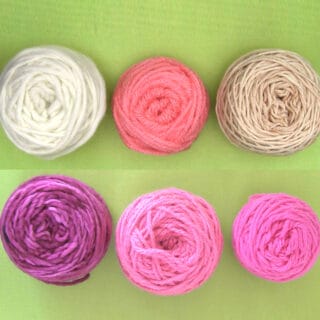 Six yarn balls in purple, pink, and white yarn colors arranged atop a green background.