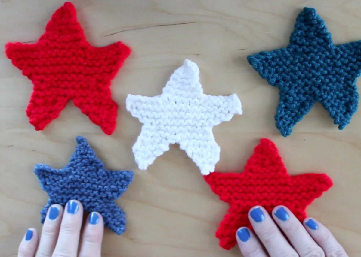 5 knitted star shapes on a tabletop with woman's hands in red, white, and blue yarn colors.