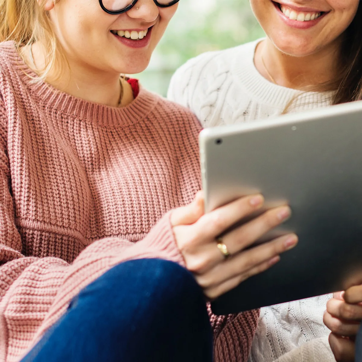 Two women looking at a digital tablet wearing knitted sweaters while smiling.