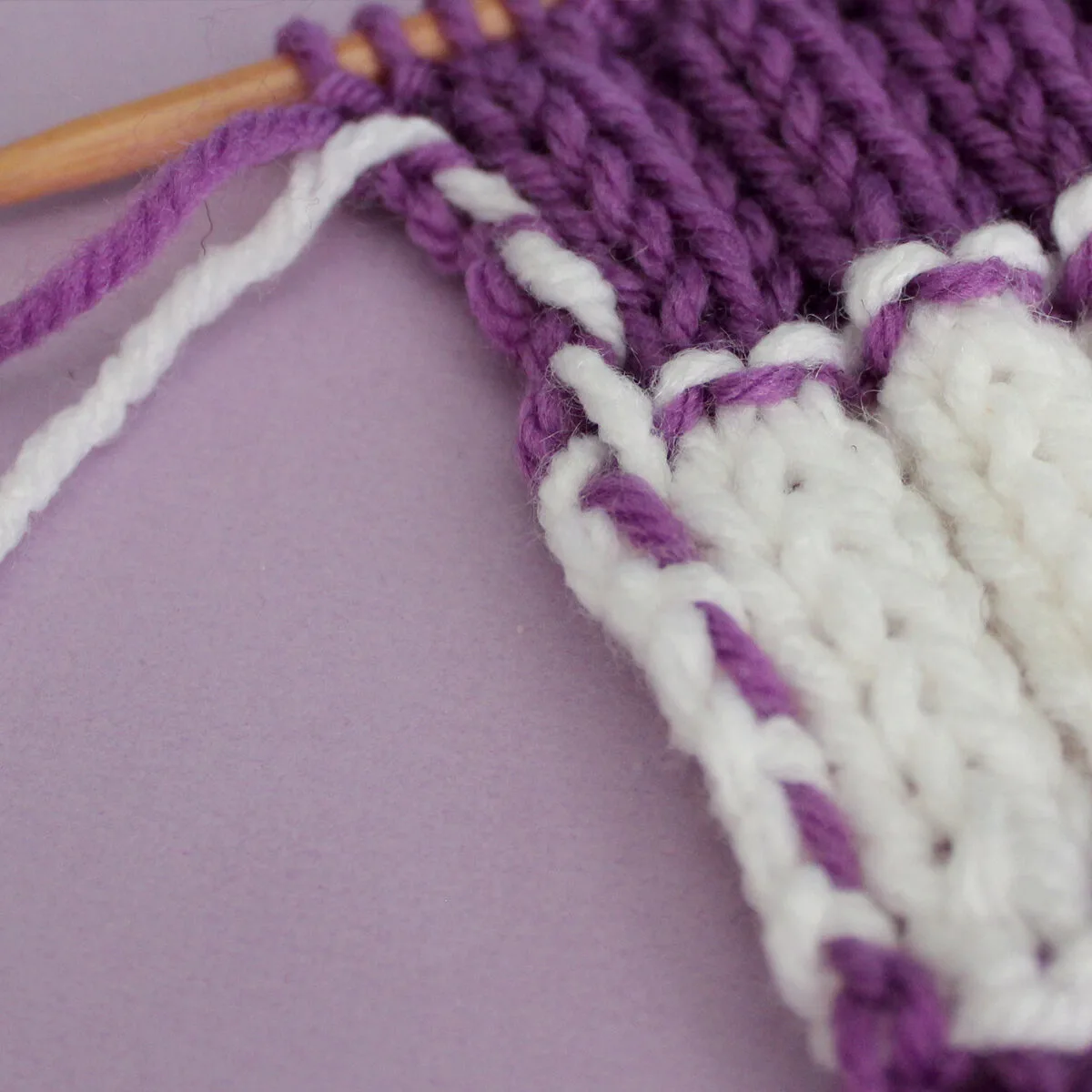 Yarn Carried up the side of a knitted swatch in stripes of purple and white colored yarn.
