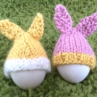 Two eggs with knitted easter bunny rabbit ear toppers as cozies on a green backdrop.