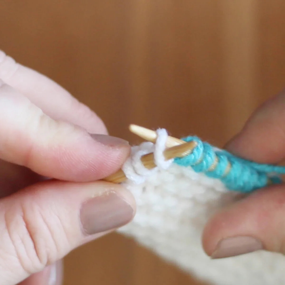 Two hands holding knitting needles demonstrating the Knit Front and Back technique.