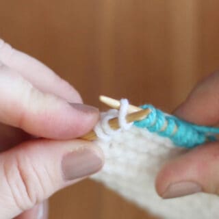 Two hands holding knitting needles demonstrating the Knit Front and Back technique.