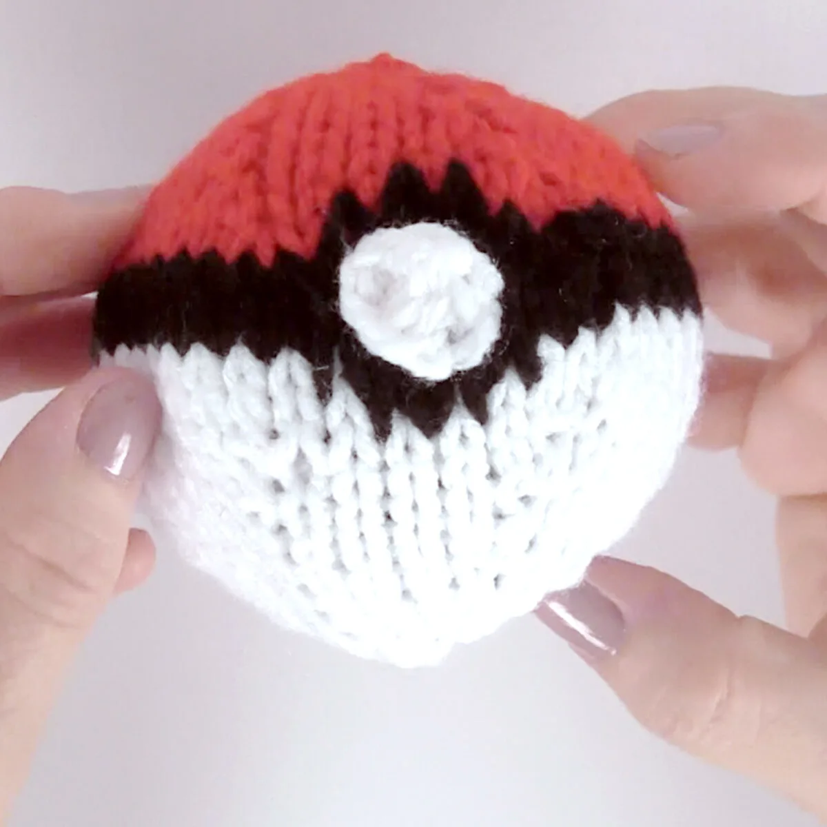 Knitted Poke Ball in white, red, and black yarn colors held by two hands.