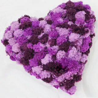 Knitted heart shaped pillow in pom pom yarn in shades of purple yarn color.