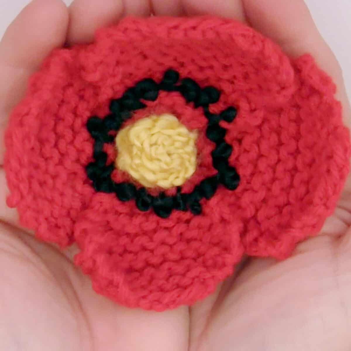 Knitted Poppy Flower in red color yarn held by two hands.