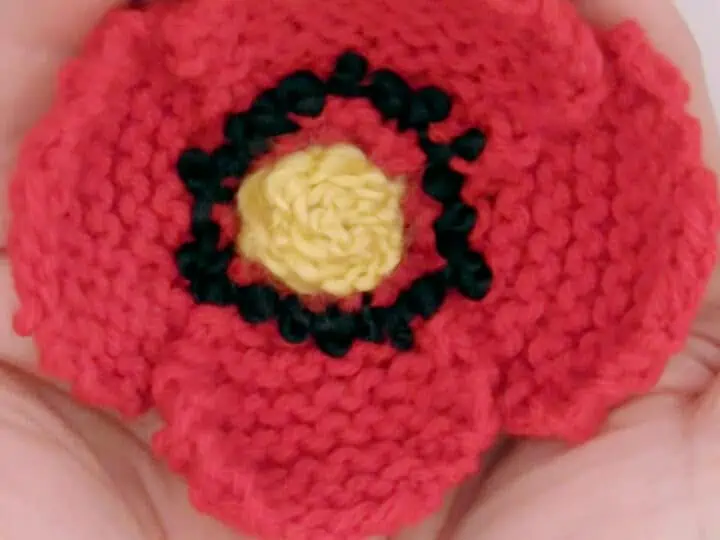 Knitted Poppy Flower in red color yarn held by two hands.
