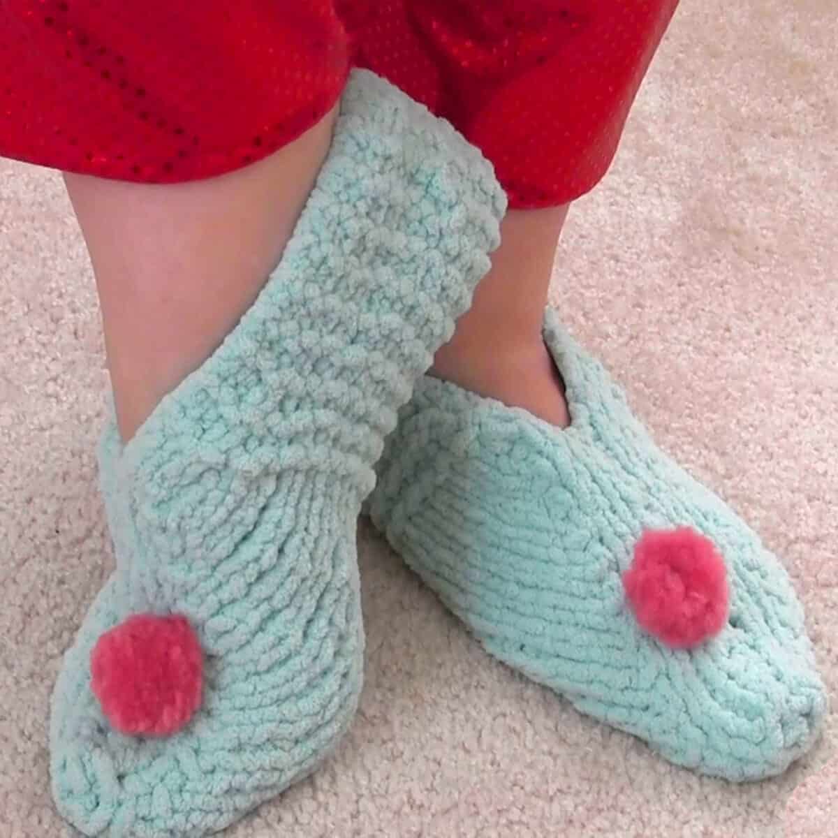 Knitted slippers in light blue yarn color with pink pom poms.