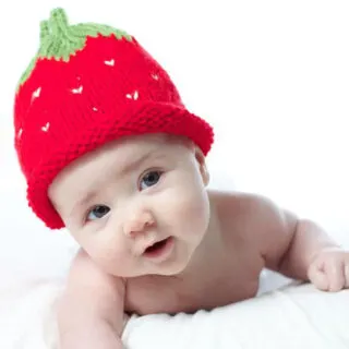 Baby wearing a knitted strawberry hat in red, white, and green yarn colors.