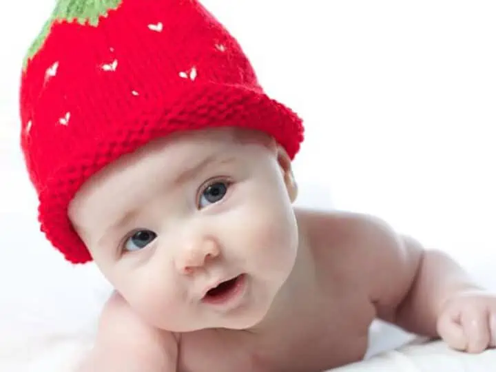 Baby wearing a knitted strawberry hat in red, white, and green yarn colors.